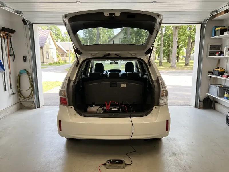 White hybrid in a garage with its tailgate open and smart charger connected to the auxiliary battery beneath the boot floor; the garage door stays open throughout.