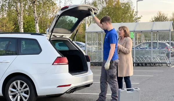 Technician holding open the boot of a white estate car in a shopping-centre car park; boot-opening tools travel in a separate case.