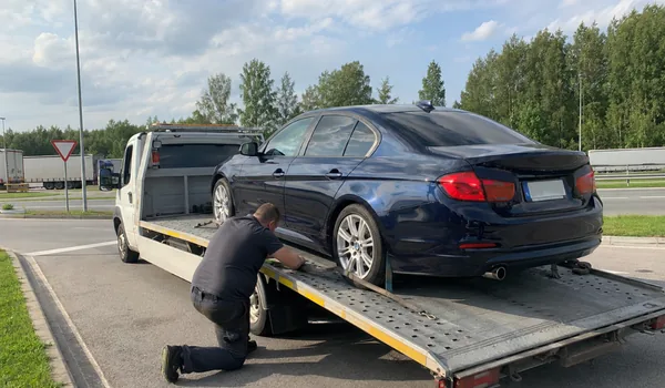 Auto transport linnade vahel, tehnik kinnitab tumesinise sõiduauto platvormveokile maantee ääres, rihma vaba ots seotakse kinni, et see maanteekiirusel ei lehviks.
