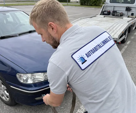 Tehnik kinnitab tumesinise sõiduauto esiosale rihma, taga langetatud platvormiga puksiirauto, vintsi konksu turvalukk suletakse enne tõmbe algust.