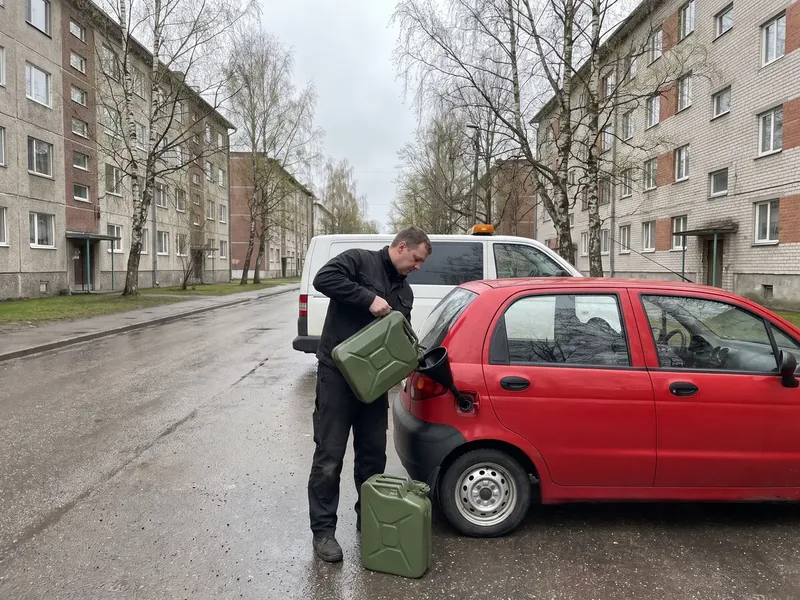 Tehnik valab Kallavere kortermajade vahel märjal tänaval rohelisest kanistrist lehtri kaudu kütust punase linnaauto paaki, maas teine kanister ja taga kollase vilkuriga kaubik, pärast valamist sõidab tehnik kaubikuga kliendi järel lähima tanklani.
